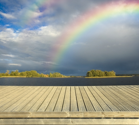 Empty wooden jetty on the lake shore with dark sky and rainbow in the backgroundの写真素材
