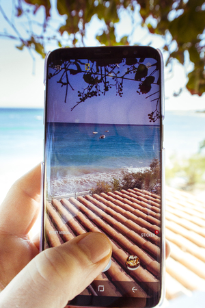 Close up hand holding smartphone taking sea view photo while sitting on beach chair on holidayの写真素材