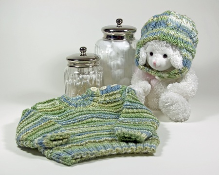 A babys hand knit sweater and matching hat on a toy lamb along with cotton balls and Q-tips in apothecary jarsの写真素材