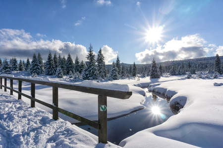 Winter mountain landscape with stream and wooden bridge. Trees and the sun are reflected in the water. Izera Mountains, Czech Republic, Europe.の写真素材