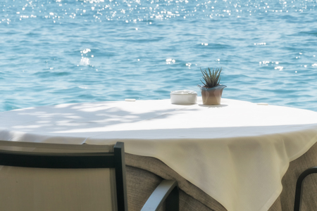 Empty table in a cafe on the shore of the sea or lake, covered with a white tablecloth with an ashtray and a decorative flower pot on a sunny summer dayの写真素材