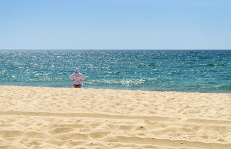 Mature woman in white shirt and white hat is standing on the beach and watching the sea in sunny summer day.の写真素材
