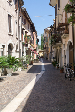 BARDOLINO, VENETO REGION, ITALY - AUGUST 7, 2017: narrow street with restaurants and shops with colorful facades of tenements and blue sky, cypress tree in background.のeditorial素材