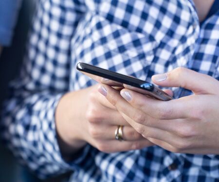 Young woman dressed in blue checkered shirt,  using modern smartphone device, female hands typing text message via cellphone, social networking concept. Selective focus.の写真素材
