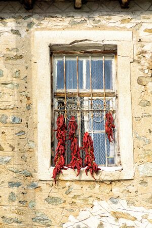 Bunches of red peppers dried in the sun hang in front of the window of old rustical building.の写真素材