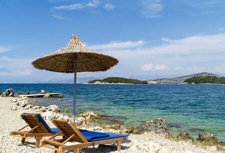 Rocky beach on the shore of the Ionian Sea with sunbeds and straw umbrellas. Blue and tourquse sea in background, nobody. Sunny Summer day. Ksamil, Albania, Europe.の写真素材