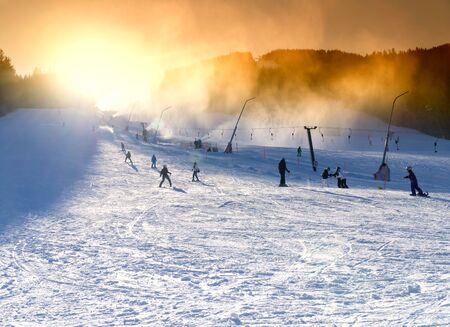 Ski resort, snowmaking on artificial slopes at sunset. Skiers and sheer snowboarders on the slopeの写真素材