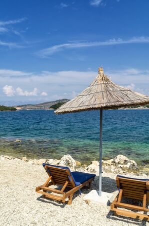 Rocky beach on the shore of the Ionian Sea with sunbeds and straw umbrellas. Blue and tourquse sea in background, nobody. Sunny Summer day. Ksamil, Albania, Europe.の写真素材
