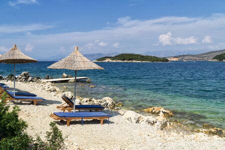 Rocky beach in Ksamil on the shore of the Ionian Sea with sunbeds and straw umbrellas. Blue and tourquse sea in background, nobody. Sunny Summer day. Ksamil, Albania, Europe.の写真素材