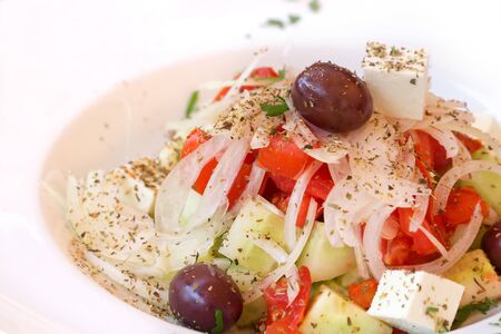 Fresh spring salad with tomato, cucumber, onion, black olives, feta cheese, olive oil, sprinkled with herbs, served on white plate with copy space.の写真素材