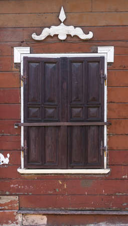 Traditional architecture detail. Brown wooden wall with window closed with wooden shutters with metal fittings and white decorative details.の写真素材