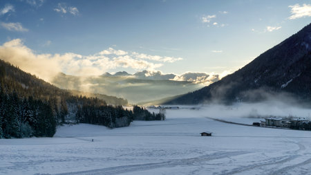 Mountain valley shrouded in mist at sunset in winter. Thick mist envelops the valley at dusk, mountains picks on horizon, clouds on blue sky. South Tyrol, Trentino Alto Adige, Alps, Italy, Europe.の写真素材