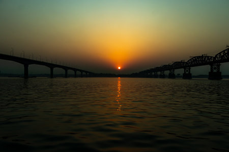 The setting sun acts as a focal point, centered between the dark of two river bridges. The hazy sky reflects a path of light across the waterの写真素材