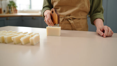 A person wearing an apron is pictured using a wooden tool to cut bars of handmade soap on a clean white countertop The craftsperson is in the process of creating soap from scratch showcasing the artisan process involved This image captures the essence of handcrafted goods and the dedication that goes into producing quality homemade products The soap bars are arranged neatly representing cleanliness and care The neutral color palette emphasizes the purity and natural ingredients of the handmade soap highlighting the beauty of the soap making processの素材