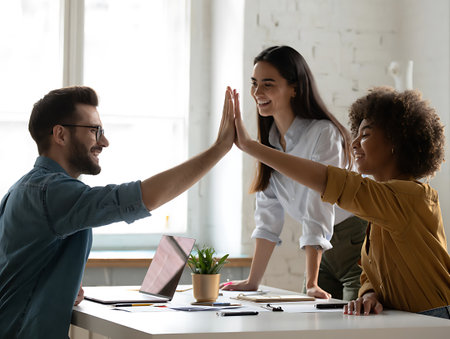 Three colleagues highfive in a bright office meeting room with a laptop and plant on the tableの素材
