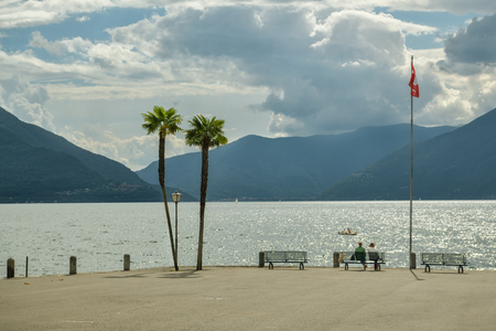 Ascona, Switzerland - September 2, 2018: Two people sitting on the bench on Lago Maggiore shore in Ascona, Switzerland during windy summer day 2018のeditorial素材