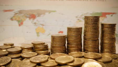 A dense arrangement of gold coins fills the foreground of with several stacks of varying heights extending into the mid-ground. Behind the coins, a map is visible, slightly out of focus, providing a global perspective. The lighting emphasizes the golden hue and reflective surfaces of the coins, creating a sense of abundance and wealth. The conveys a strong theme of financial prosperity and the global nature of economic activity.の素材