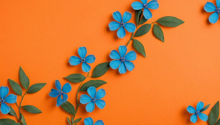 A graceful, flowing arrangement of blue flowers and green leaves is depicted against a bright orange background. The are arranged as if growing along a winding branch or vine, creating a dynamic and organic visual path across the frame. The blue flowers, with their five petals, are interspersed with clusters of vibrant green leaves. The composition feels natural and with the cool tones of the flowers and leaves providing a contrast to the warm, energetic orange surface.の素材
