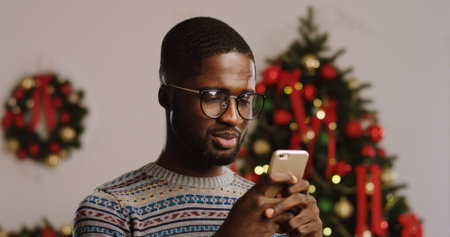 Close up of young African American man in glasses taping on his smart phone and smiling. Christmas...