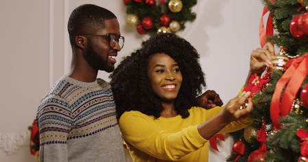 Close up of young attractive smiling African American couple preparing for the Christmas and decorating the Christmas tree with red ribbons and golden baubles. Inside the living room. Portrait shotの写真素材