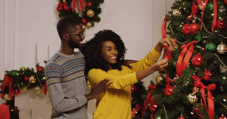 Young happy african american couple decorating the Christmas tree with nice Christmas baubles in the cozy living room. Portraitの写真素材