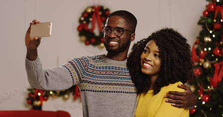 Portrait shot of young smiled and happy African American couple having videochat on a smart phone in the cozy decorated living room. Christmas tree with lights and red ribbons on the background. Indoorの写真素材