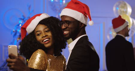 Portrait of young happy African American couple in santa clause hats taking selfies on a smart phone at the New Year or Christmas party. Dancing people background. Indoorの写真素材
