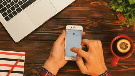 New York - September 10, 2017: Man using calendar app on a white smartphone. Man making plans for future, making a new event. Top view on the old wooden table with cup of tea, notebook and laptop computer.のeditorial素材