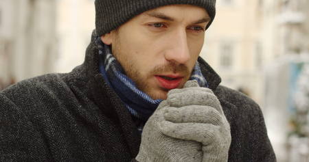 Close up of the sad handsome man in a hat and warm clothes thinking and warming his hands in gloves on the street with Christmas decorations. Portrait. Outdoorの写真素材
