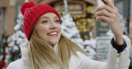 Close up of the attractive blonde woman videochating on the smartphone while standing in the winter street. Christmas trees and shop window on the background. Portrait shot. Outdoorの写真素材