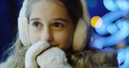 Portrait of the charming blonde girl warming her hands in the white gloves and dreaming about something in the evening. Christmas illuminations on the blurred background. Outside. Close upの写真素材