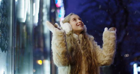 Happy pretty girl with long blonde hair spinning around with her hands up while it is snowing near shop showcase in the winter evening. Outdoor.の写真素材