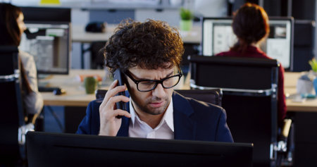 Portrait of angry handsome male office worker in glasses talking on the phone while working on the...