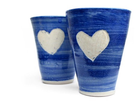 Two ceramic handcrafted blue mugs with white heart shape on each, focus on front one, isolated on whiteの写真素材