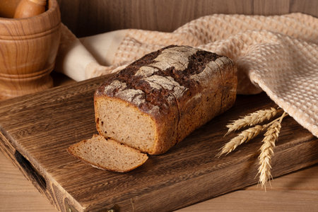 Freshly baked whole grain bread resting on a wooden cutting board with woven cloth and wheat stalks...