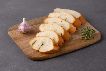 Freshly sliced bread with garlic and herbs displayed on a wooden cutting board on grey table
