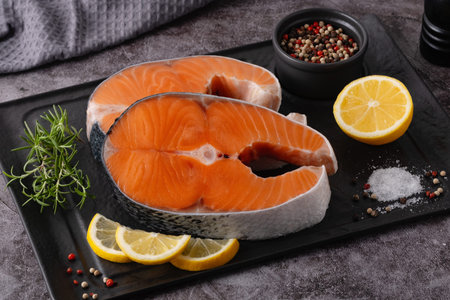 Two salmon fillets are placed on a black plate with lemon slices. A small bowl of mixed pepper and salt sit nearby. The setting looks like a kitchen area prepared for cooking.の写真素材