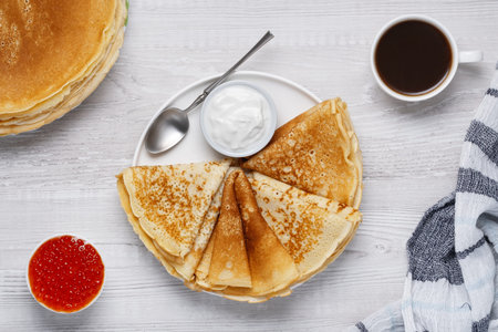 Pancakes are served with sour cream and red caviar on a wooden table, accompanied by coffee