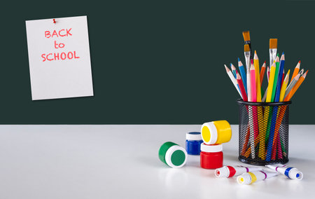 Pencil's holder with colourful pencils and paint brushes, colourful paints on white desk and notebook sheet with text on dark green chalkboard. Back to school concept. Copy spaceの写真素材