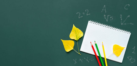 Copybook with colourful pencils and yellow leaves on dark green chalkboard flat lay. Back to school...