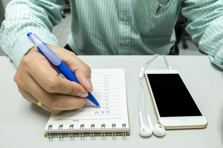 Business man use blue pen writing a to do list on notebook with smart phone and earphoneの写真素材
