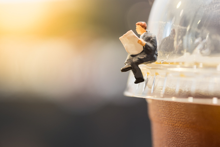 Reading, relaxing, hobby concept. Close up of businessman miniature mini figure sit and read newspaper on a take away plastic cup iced black coffee (Americano)の写真素材