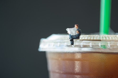 Business, reading, relaxing, hobby concept. Close up of businessman miniature mini figure sit and read newspaper on a take away plastic cup iced black coffee (Americano)の写真素材