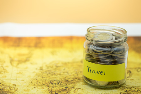Time, Travel and Saving Concept. Full of coins in clear bottle with yellow label note with Travel word on wolrd map.の写真素材