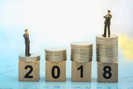 2018 New year business and saving concept. Close up of two businessman miniature people figures standing and looking to the top on stack of silver coins  on number wooden block on world map.の写真素材