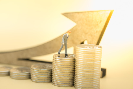 Money, business, saving and growth concept. Close up of businessman miniature figure walking on top of stack of silver coins with wooden arrow symbol.の写真素材