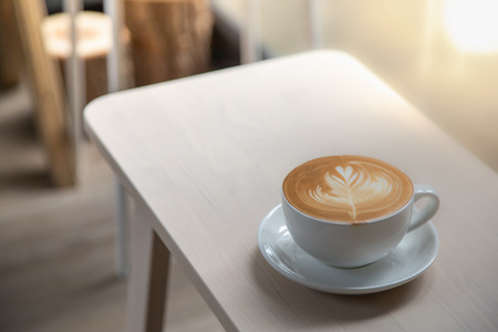 Close up of white cup of hot coffee latte with milk foam heart and flower shape art on wooden table.の写真素材