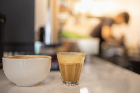 Close up of shot glass and white cup of hot coffee latte with coffee maker machine and barista as background.の写真素材