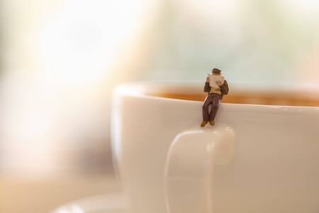 Businessman miniature figure sitting and reading a newspaper on white cup of hot coffee with copy scape.の写真素材