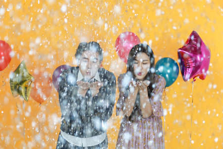 Holiday Celebrate, Christmas and New year party concept. Portrait of asian man and woman blowing silver paper confetti with colorful balloon and yellow background.の写真素材
